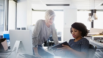 Two women at a desk looking at a tablet and discussing content on it. 